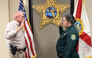 Agency photo of Sheriff Kitchings with hand raised swearing in new Detention Deputy Victoria Chatigny. Both in uniform with agency Sheriff star in center flanked by the US flag and the Florida state flag.