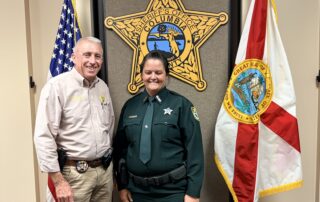 Sheriff Kitchings and Detention Deputy Chatigny facing family after swearing in ceremony flanked by US flag and Florida state flag.